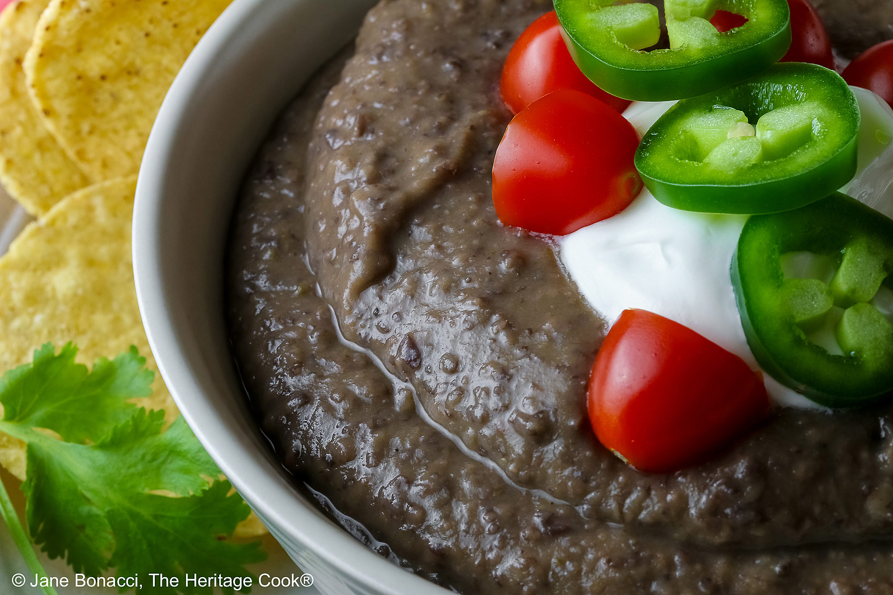 Chipotle Black Bean Soup in a pale blue bowl sitting on a white plate with tan rim; cilantro, tomatoes, lime slices, tortilla chips around the bowl on the edge of the plate; soup topped with sour cream, cherry tomatoes, and sliced jalapeno pepper © 2025 Jane Bonacci, The Heritage Cook.