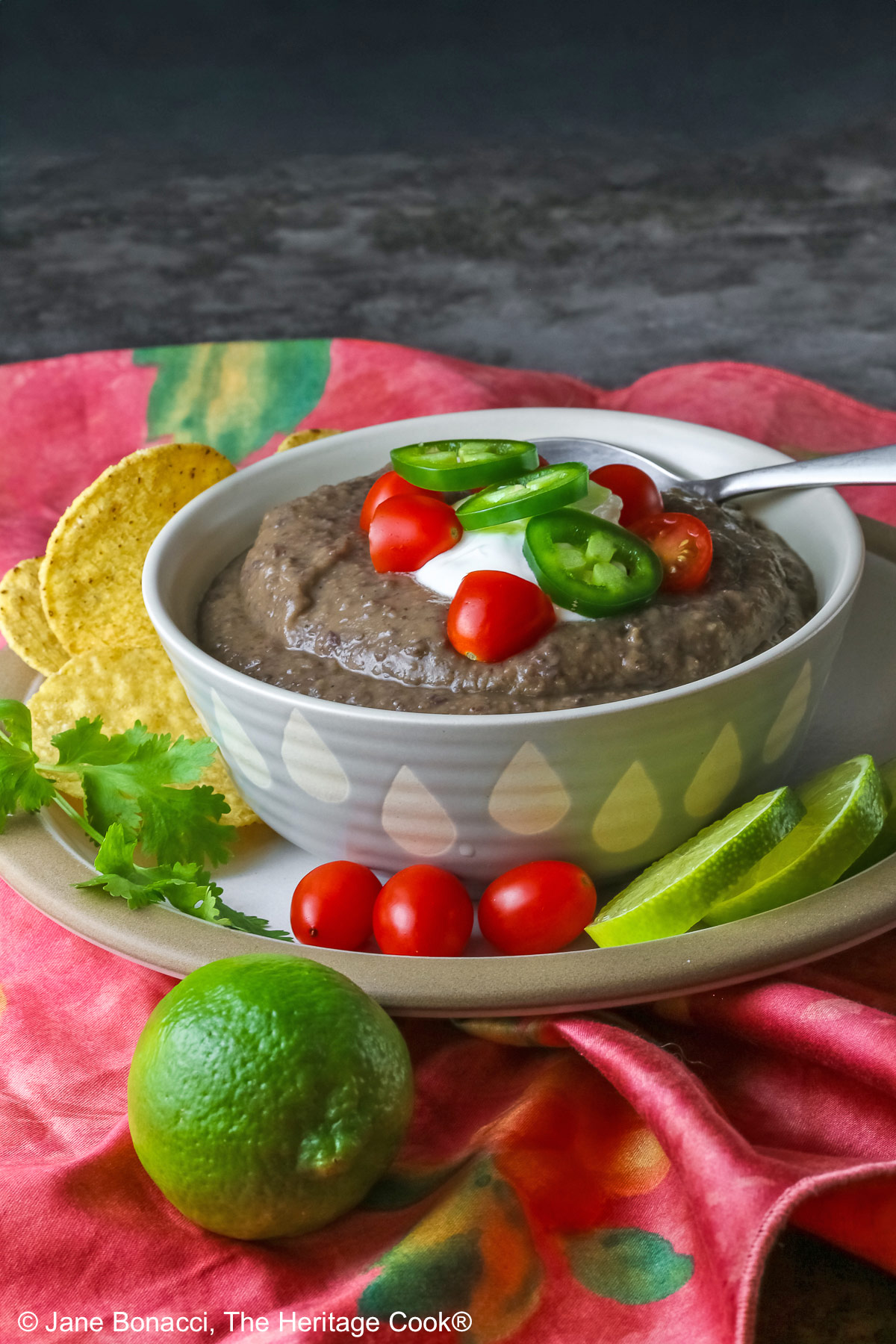 Chipotle Black Bean Soup in a pale blue bowl sitting on a white plate with tan rim; cilantro, tomatoes, lime slices, tortilla chips around the bowl on the edge of the plate; soup topped with sour cream, cherry tomatoes, and sliced jalapeno pepper © 2025 Jane Bonacci, The Heritage Cook.