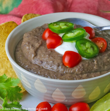 Chipotle Black Bean Soup in a pale blue bowl sitting on a white plate with tan rim; cilantro, tomatoes, lime slices, tortilla chips around the bowl on the edge of the plate; soup topped with sour cream, cherry tomatoes, and sliced jalapeno pepper © 2025 Jane Bonacci, The Heritage Cook.