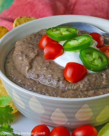 Chipotle Black Bean Soup in a pale blue bowl sitting on a white plate with tan rim; cilantro, tomatoes, lime slices, tortilla chips around the bowl on the edge of the plate; soup topped with sour cream, cherry tomatoes, and sliced jalapeno pepper © 2025 Jane Bonacci, The Heritage Cook.