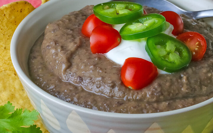 Chipotle Black Bean Soup in a pale blue bowl sitting on a white plate with tan rim; cilantro, tomatoes, lime slices, tortilla chips around the bowl on the edge of the plate; soup topped with sour cream, cherry tomatoes, and sliced jalapeno pepper © 2025 Jane Bonacci, The Heritage Cook.