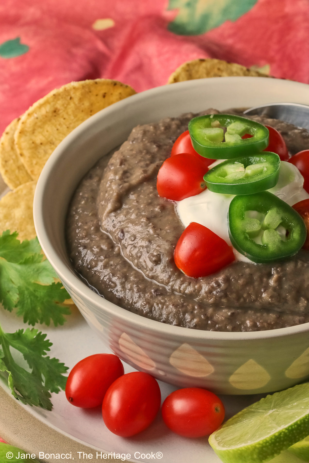 Chipotle Black Bean Soup in a pale blue bowl sitting on a white plate with tan rim; cilantro, tomatoes, lime slices, tortilla chips around the bowl on the edge of the plate; soup topped with sour cream, cherry tomatoes, and sliced jalapeno pepper © 2025 Jane Bonacci, The Heritage Cook.
