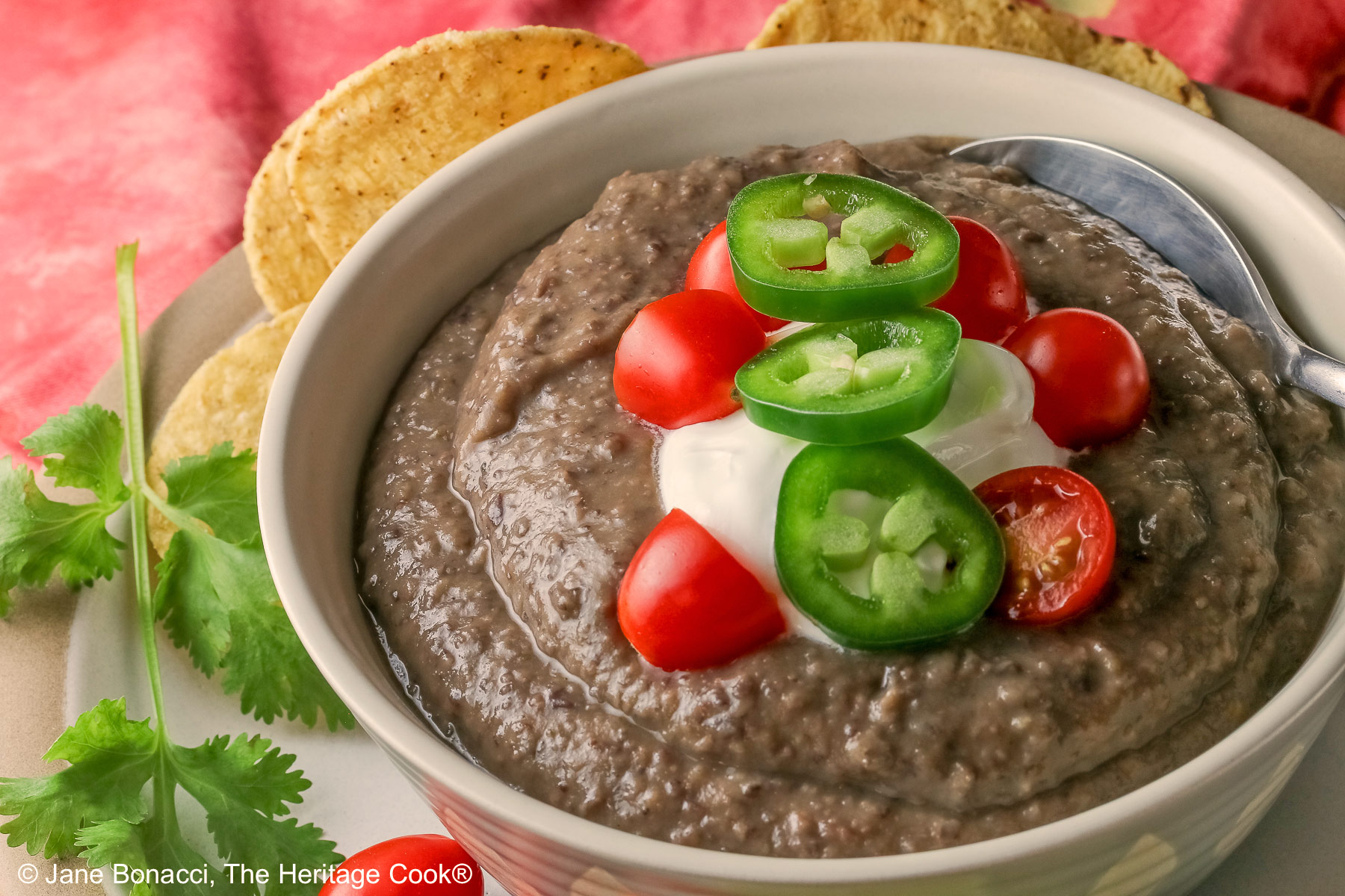 Chipotle Black Bean Soup in a pale blue bowl sitting on a white plate with tan rim; cilantro, tomatoes, lime slices, tortilla chips around the bowl on the edge of the plate; soup topped with sour cream, cherry tomatoes, and sliced jalapeno pepper © 2025 Jane Bonacci, The Heritage Cook.