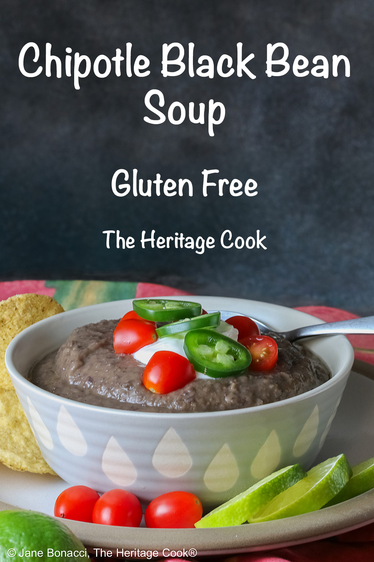 Chipotle Black Bean Soup in a pale blue bowl sitting on a white plate with tan rim; cilantro, tomatoes, lime slices, tortilla chips around the bowl on the edge of the plate; soup topped with sour cream, cherry tomatoes, and sliced jalapeno pepper © 2025 Jane Bonacci, The Heritage Cook.