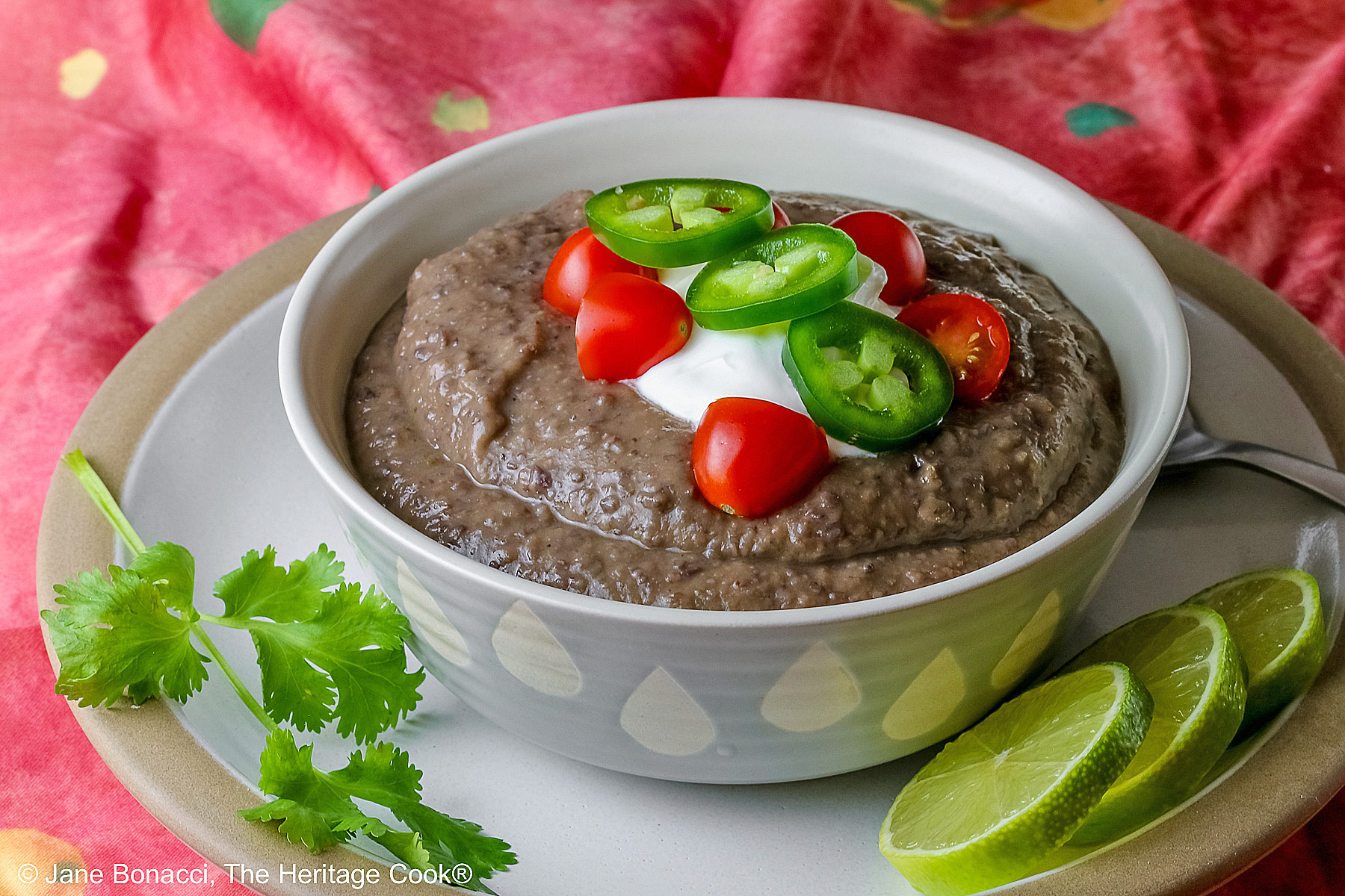Chipotle Black Bean Soup in a pale blue bowl sitting on a white plate with tan rim; cilantro, tomatoes, lime slices, tortilla chips around the bowl on the edge of the plate; soup topped with sour cream, cherry tomatoes, and sliced jalapeno pepper © 2025 Jane Bonacci, The Heritage Cook.
