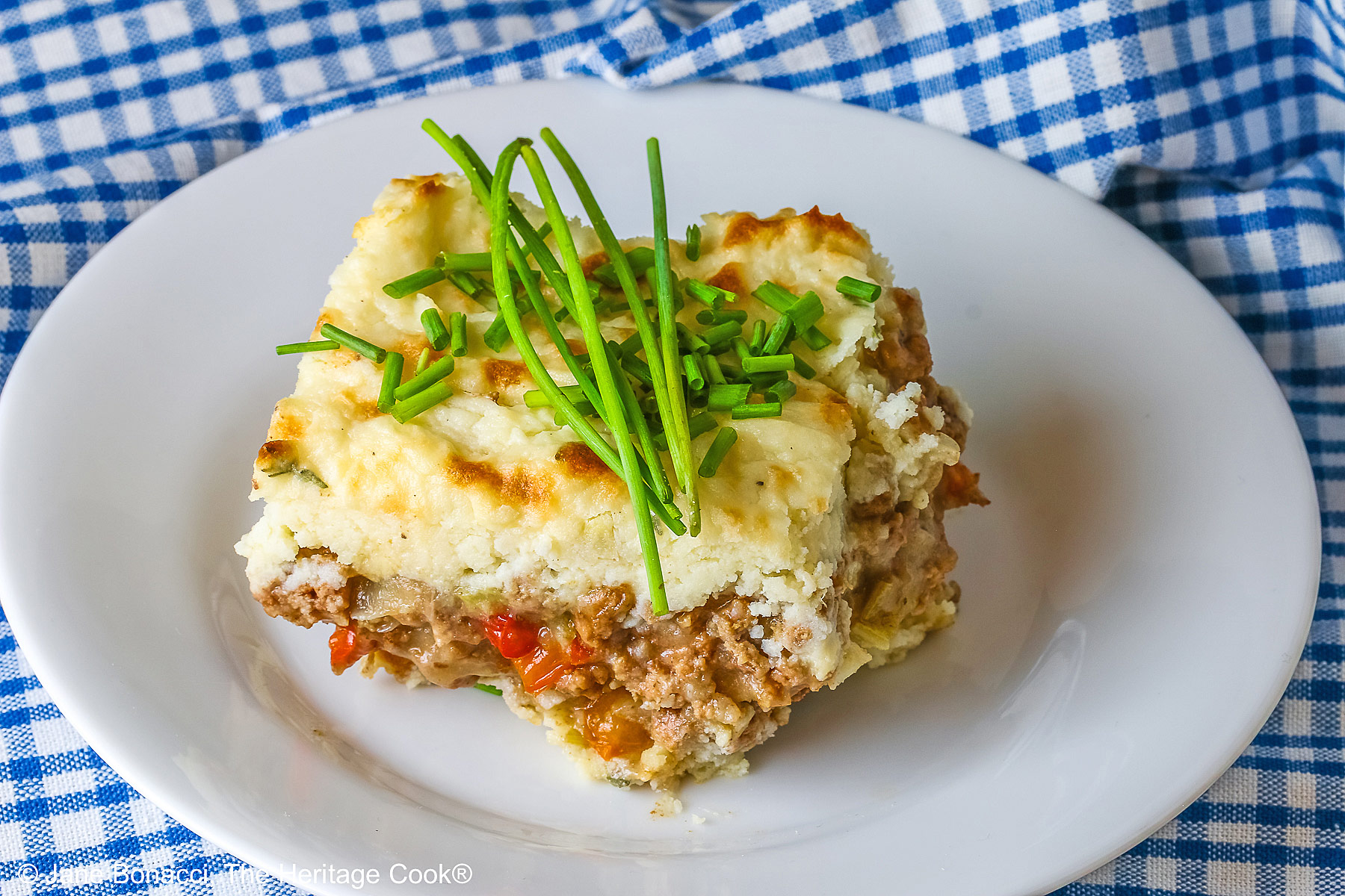 Square serving of Beef Pie with Garlic-Thyme Mashed Potatoes on a white plate, sitting on a blue checked cloth sprinkled with chopped chives and whole chives laid across the top, most with a fork nestled on the edge © 2026 Jane Bonacci, The Heritage Cook.