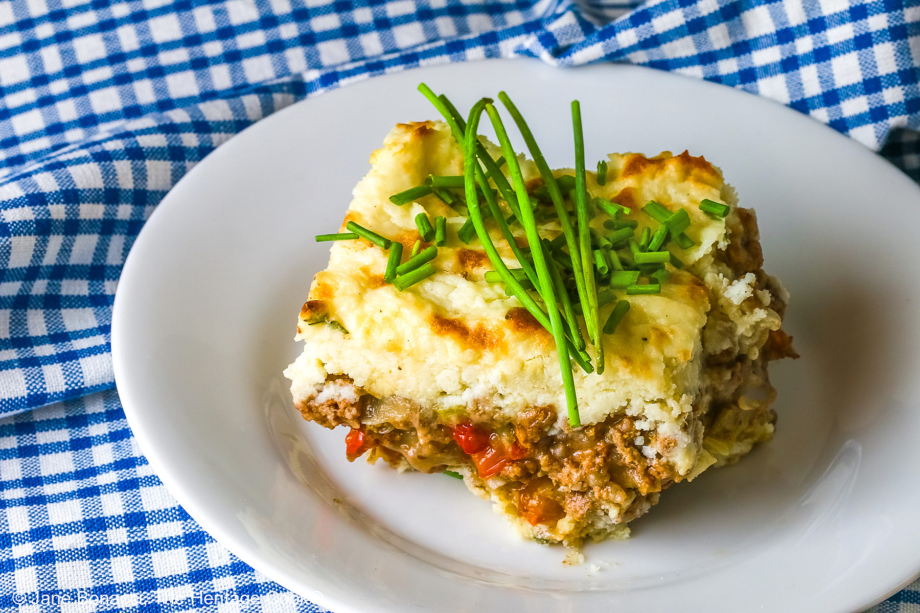 Square serving of Beef Pie with Garlic-Thyme Mashed Potatoes on a white plate, sitting on a blue checked cloth sprinkled with chopped chives and whole chives laid across the top, most with a fork nestled on the edge © 2026 Jane Bonacci, The Heritage Cook.