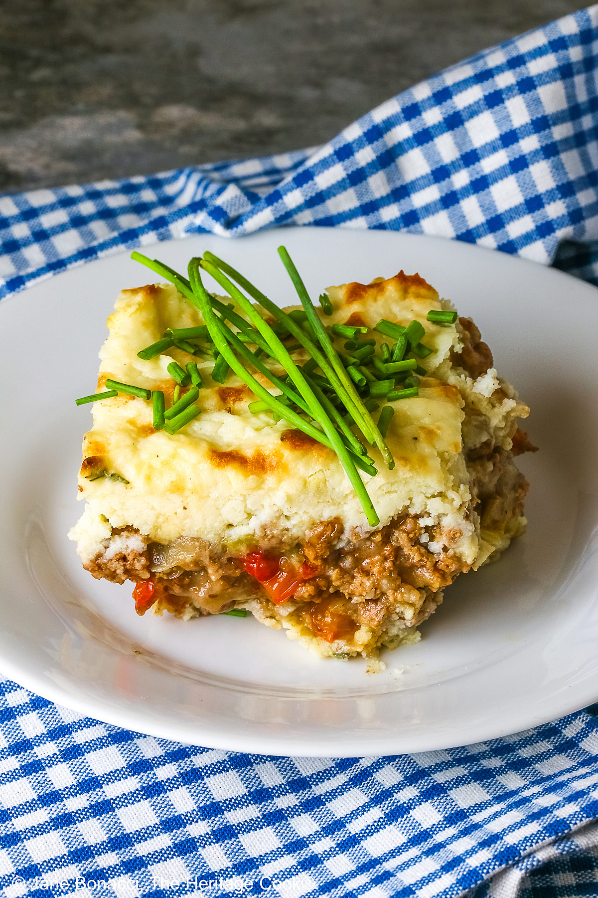 Square serving of Beef Pie with Garlic-Thyme Mashed Potatoes on a white plate, sitting on a blue checked cloth sprinkled with chopped chives and whole chives laid across the top, most with a fork nestled on the edge © 2026 Jane Bonacci, The Heritage Cook.