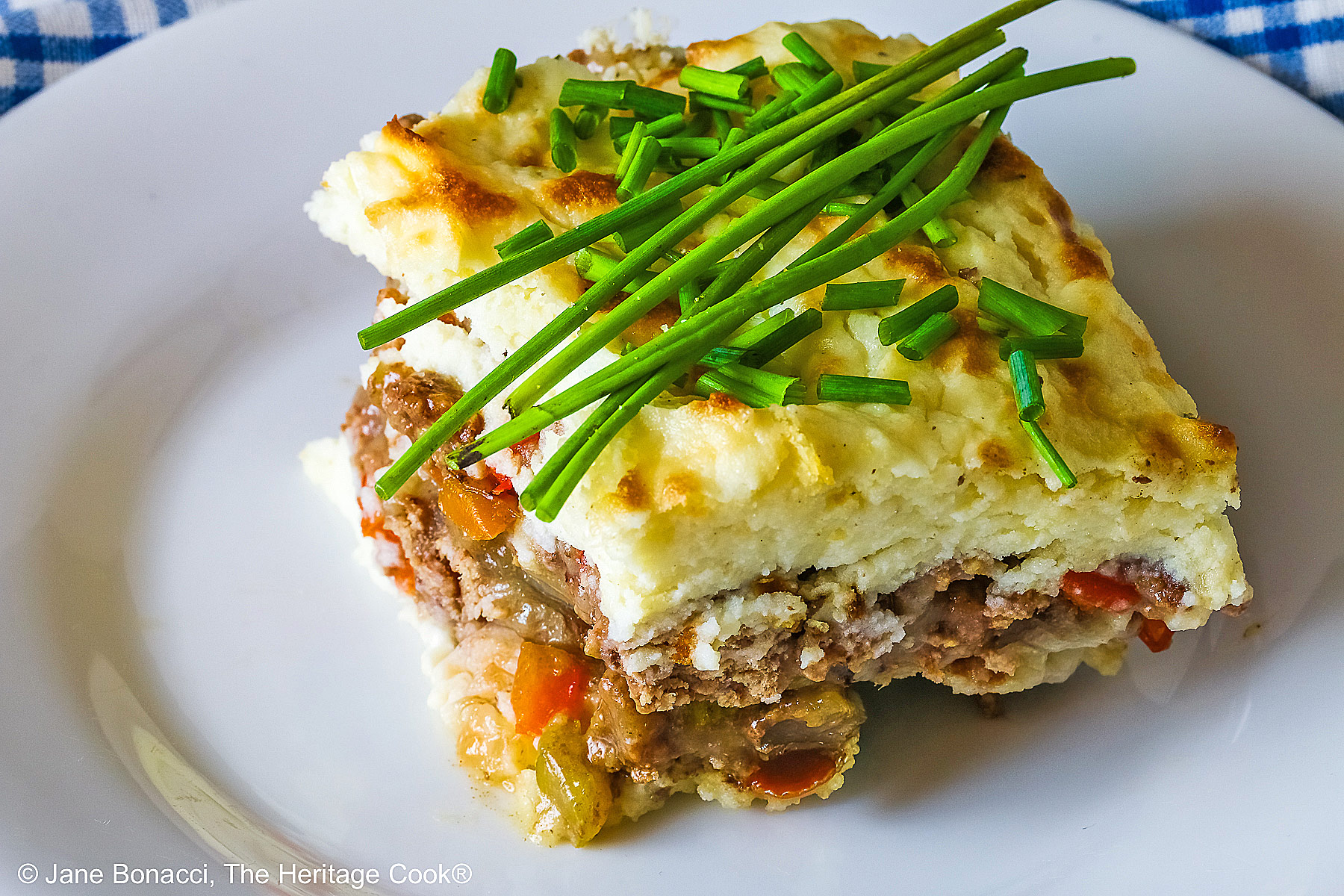 Square serving of Beef Pie with Garlic-Thyme Mashed Potatoes on a white plate, sitting on a blue checked cloth sprinkled with chopped chives and whole chives laid across the top, most with a fork nestled on the edge © 2026 Jane Bonacci, The Heritage Cook.