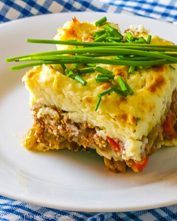 Square serving of Beef Pie with Garlic-Thyme Mashed Potatoes on a white plate, sitting on a blue checked cloth sprinkled with chopped chives and whole chives laid across the top, most with a fork nestled on the edge © 2026 Jane Bonacci, The Heritage Cook.