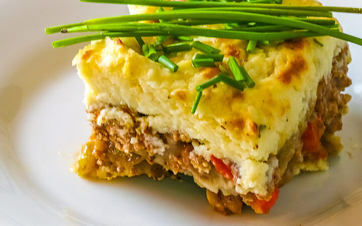 Square serving of Beef Pie with Garlic-Thyme Mashed Potatoes on a white plate, sitting on a blue checked cloth sprinkled with chopped chives and whole chives laid across the top, most with a fork nestled on the edge © 2026 Jane Bonacci, The Heritage Cook.