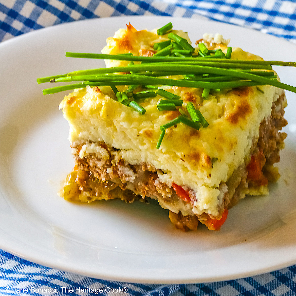 Square serving of Beef Pie with Garlic-Thyme Mashed Potatoes on a white plate, sitting on a blue checked cloth sprinkled with chopped chives and whole chives laid across the top, most with a fork nestled on the edge © 2026 Jane Bonacci, The Heritage Cook.