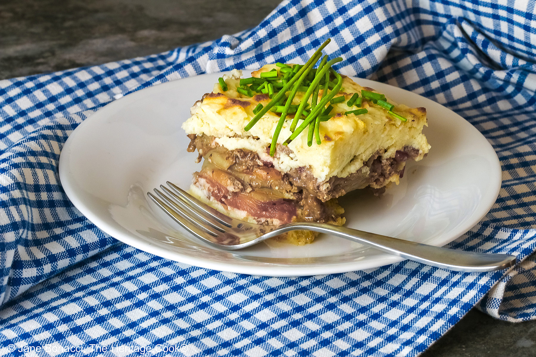 Square serving of Beef Pie with Garlic-Thyme Mashed Potatoes on a white plate, sitting on a blue checked cloth sprinkled with chopped chives and whole chives laid across the top, most with a fork nestled on the edge © 2026 Jane Bonacci, The Heritage Cook.