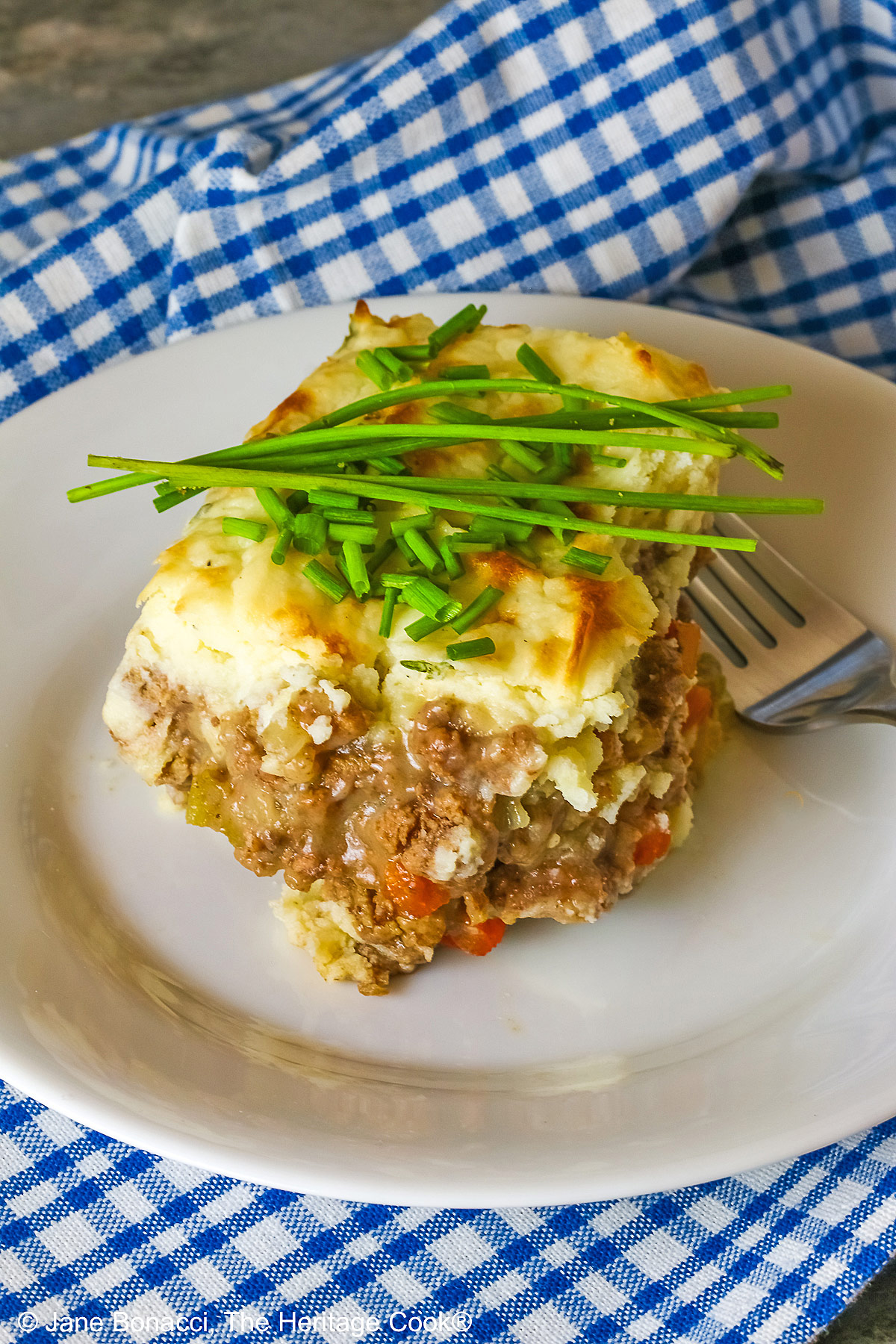 Square serving of Beef Pie with Garlic-Thyme Mashed Potatoes on a white plate, sitting on a blue checked cloth sprinkled with chopped chives and whole chives laid across the top, most with a fork nestled on the edge © 2026 Jane Bonacci, The Heritage Cook.