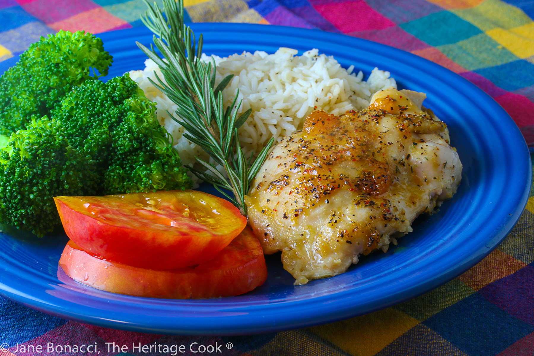 Golden chicken with an apricot-wine glaze served with sliced red tomatoes, rice, and broccoli on a blue plate placed on a colorful plaid napkin © 2023 Jane Bonacci, The Heritage Cook. 