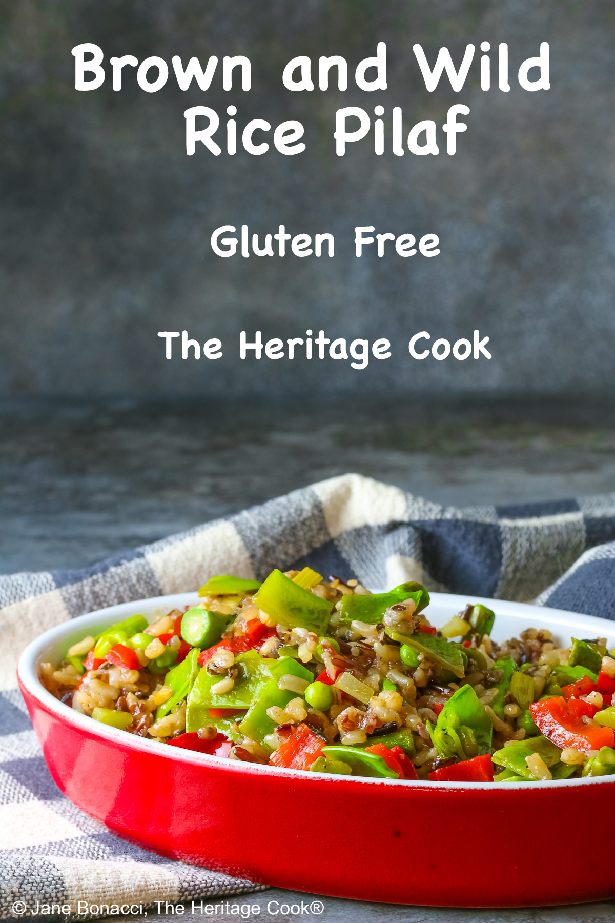 Brown & Wild Rice Pilaf piled into a red au gratin dish stuffed with lots of green vegetables and ringed with red cherry tomatoes, with a spoon tucked into the back of the dish, sitting on a grey and white checked cloth © 2025 Jane Bonacci, The Heritage Cook. 