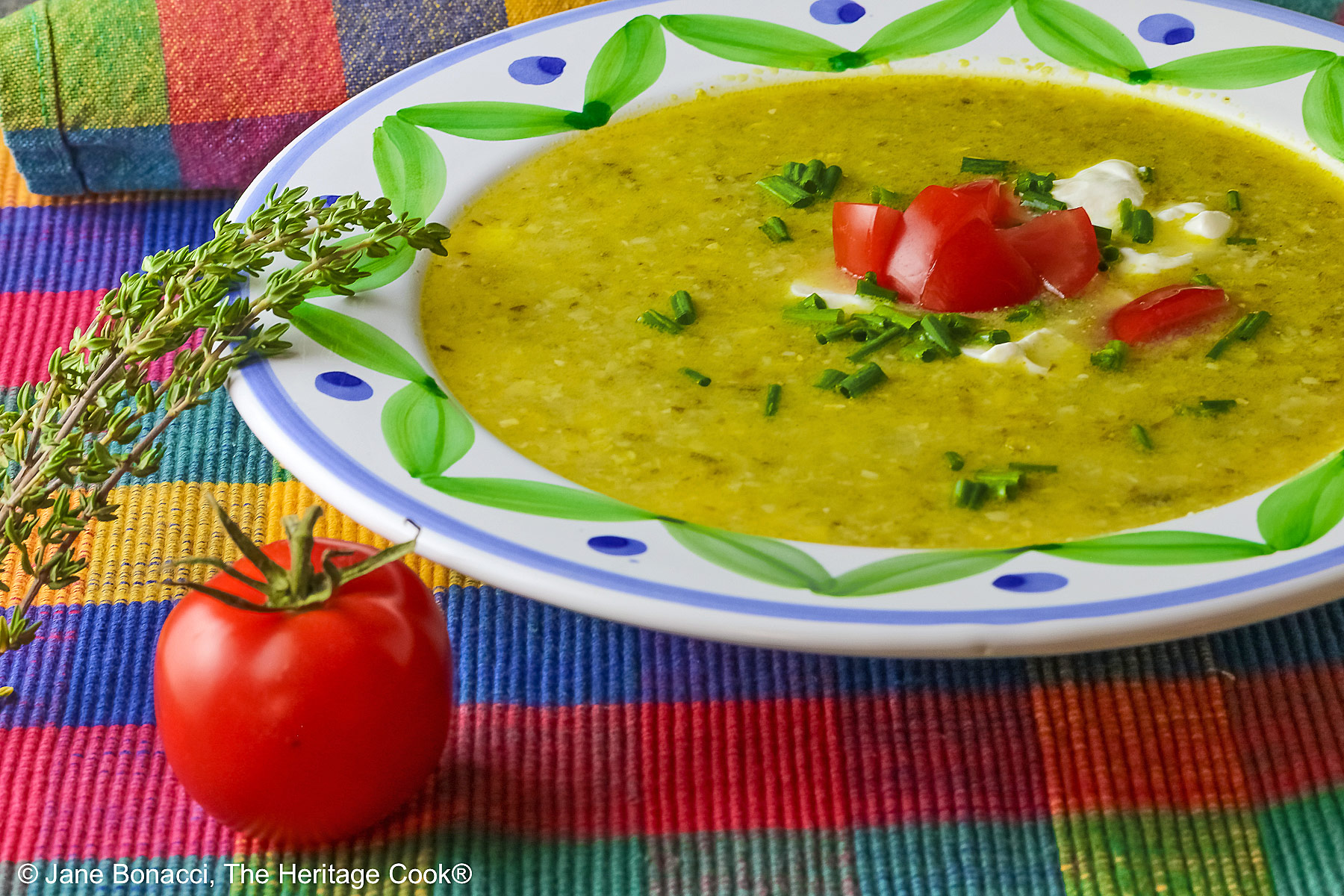 Bowl of Corn, Poblano, and Tomatillo Soup, yellowish green in color, in a bowl with green and blue decorations around the rim, topped with sour cream, chives, and chopped tomatoes, sitting on a multi-colored plaid placemat with a brightly colored cloth behind, a tomato and sprig of thyme on the left front of the bowl © 2025 Jane Bonacci, The Heritage Cook. 