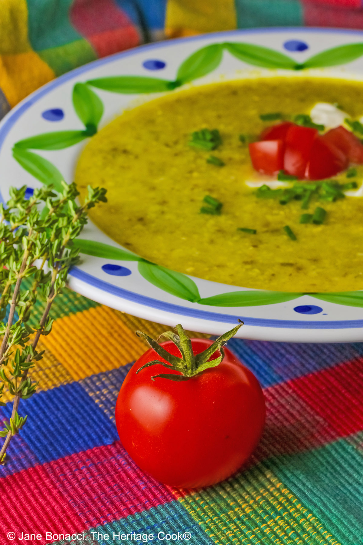 Bowl of Corn, Poblano, and Tomatillo Soup, yellowish green in color, in a bowl with green and blue decorations around the rim, topped with sour cream, chives, and chopped tomatoes, sitting on a multi-colored plaid placemat with a brightly colored cloth behind, a tomato and sprig of thyme on the left front of the bowl © 2025 Jane Bonacci, The Heritage Cook. 