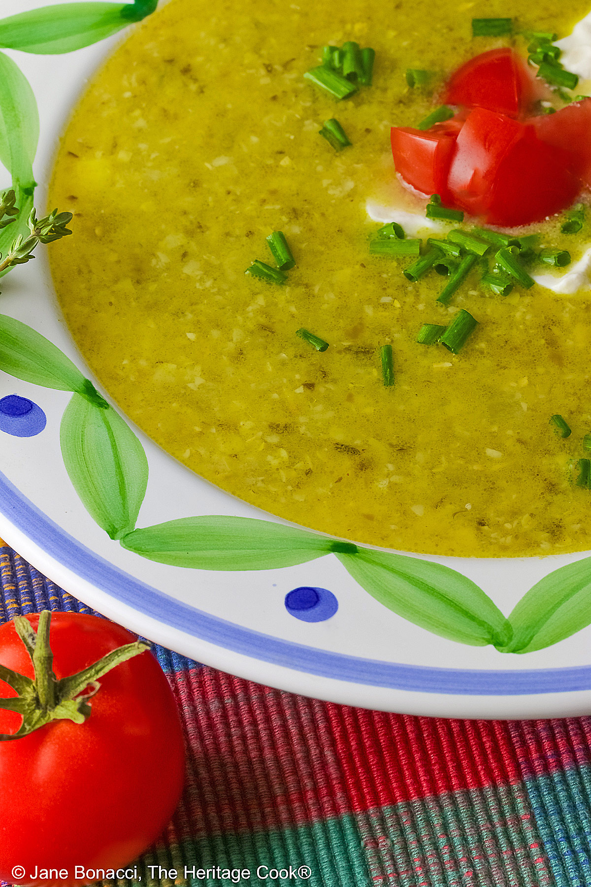 Bowl of Corn, Poblano, and Tomatillo Soup, yellowish green in color, in a bowl with green and blue decorations around the rim, topped with sour cream, chives, and chopped tomatoes, sitting on a multi-colored plaid placemat with a brightly colored cloth behind, a tomato and sprig of thyme on the left front of the bowl © 2025 Jane Bonacci, The Heritage Cook. 