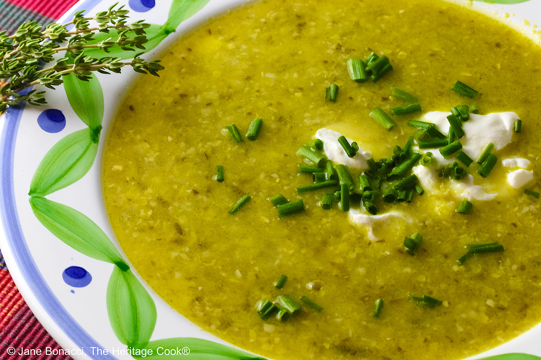 Bowl of Corn, Poblano, and Tomatillo Soup, yellowish green in color, in a bowl with green and blue decorations around the rim, topped with sour cream, chives, and chopped tomatoes, sitting on a multi-colored plaid placemat with a brightly colored cloth behind, a tomato and sprig of thyme on the left front of the bowl © 2025 Jane Bonacci, The Heritage Cook. 
