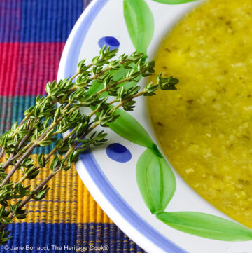Bowl of Corn, Poblano, and Tomatillo Soup, yellowish green in color, in a bowl with green and blue decorations around the rim, topped with sour cream, chives, and chopped tomatoes, sitting on a multi-colored plaid placemat with a brightly colored cloth behind, a tomato and sprig of thyme on the left front of the bowl © 2025 Jane Bonacci, The Heritage Cook.