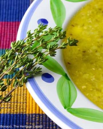 Bowl of Corn, Poblano, and Tomatillo Soup, yellowish green in color, in a bowl with green and blue decorations around the rim, topped with sour cream, chives, and chopped tomatoes, sitting on a multi-colored plaid placemat with a brightly colored cloth behind, a tomato and sprig of thyme on the left front of the bowl © 2025 Jane Bonacci, The Heritage Cook.