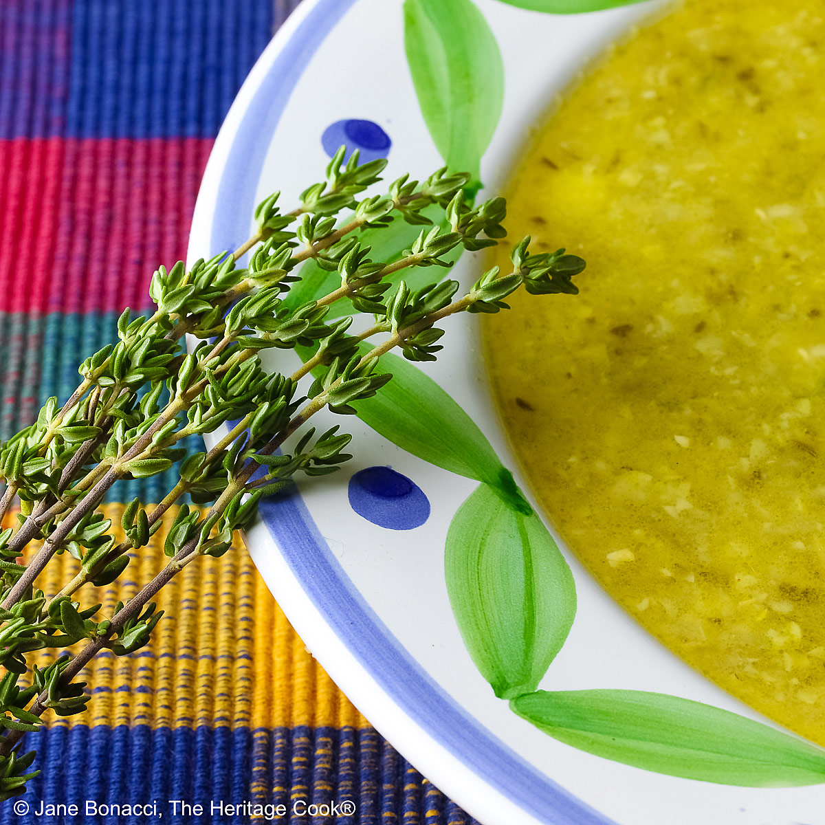 Bowl of Corn, Poblano, and Tomatillo Soup, yellowish green in color, in a bowl with green and blue decorations around the rim, topped with sour cream, chives, and chopped tomatoes, sitting on a multi-colored plaid placemat with a brightly colored cloth behind, a tomato and sprig of thyme on the left front of the bowl © 2025 Jane Bonacci, The Heritage Cook. 