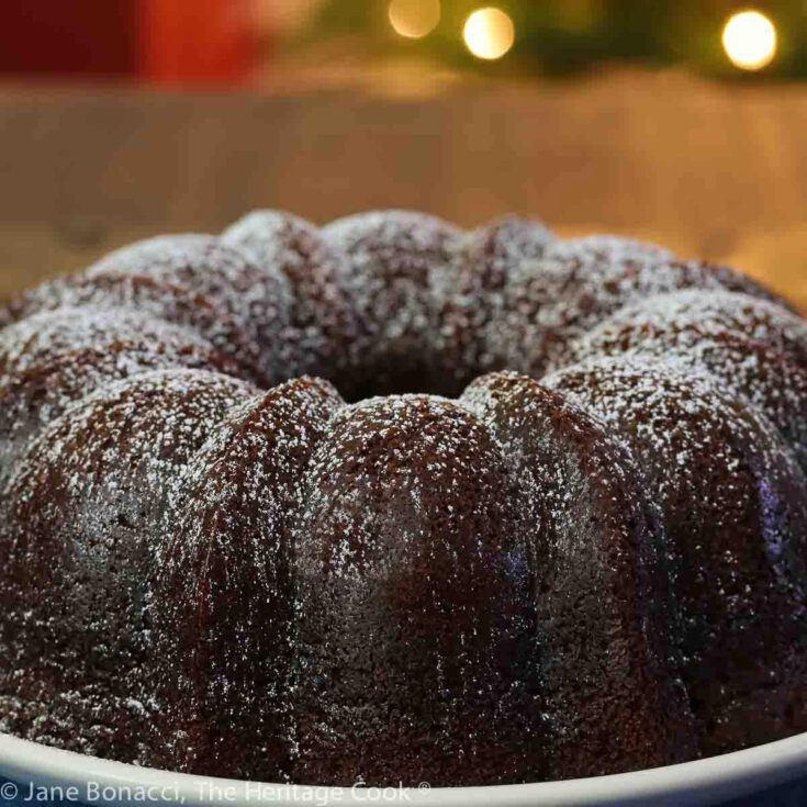 Whole Spicy Tender Gingerbread cake on a plate in front of holiday lights and placed on a blue denim cloth or a slice of the cake on a white plate with a string of painted Christmas lights around the edge © 2023 Jane Bonacci, The Heritage Cook.