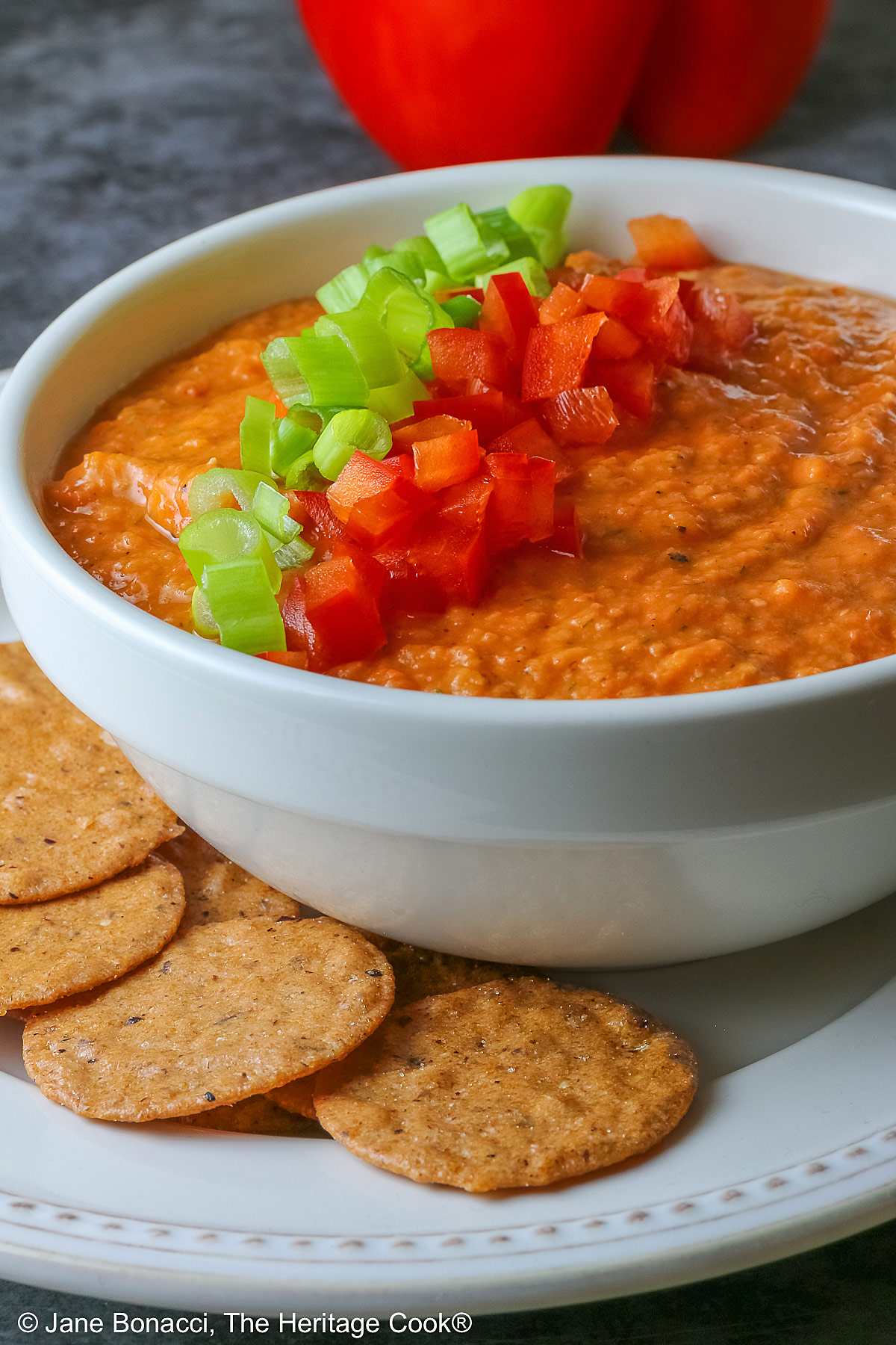 A white bowl full of White Bean and Red Bell Pepper Dip, topped with some finely chopped red bell peppers and finely sliced green onions, served with a ring of gluten-free Nut-Thin Pecan Crackers for dipping. The bowl and crackers are sitting on a cream colored plate with a red bell pepper in the back © 2026 Jane Bonacci, The Heritage Cook.
