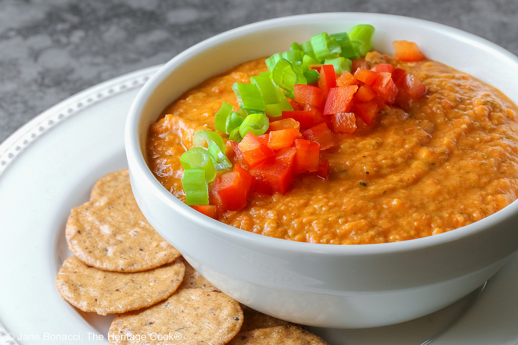A white bowl full of White Bean and Red Bell Pepper Dip, topped with some finely chopped red bell peppers and finely sliced green onions, served with a ring of gluten-free Nut-Thin Pecan Crackers for dipping. The bowl and crackers are sitting on a cream colored plate with a red bell pepper in the back © 2026 Jane Bonacci, The Heritage Cook.