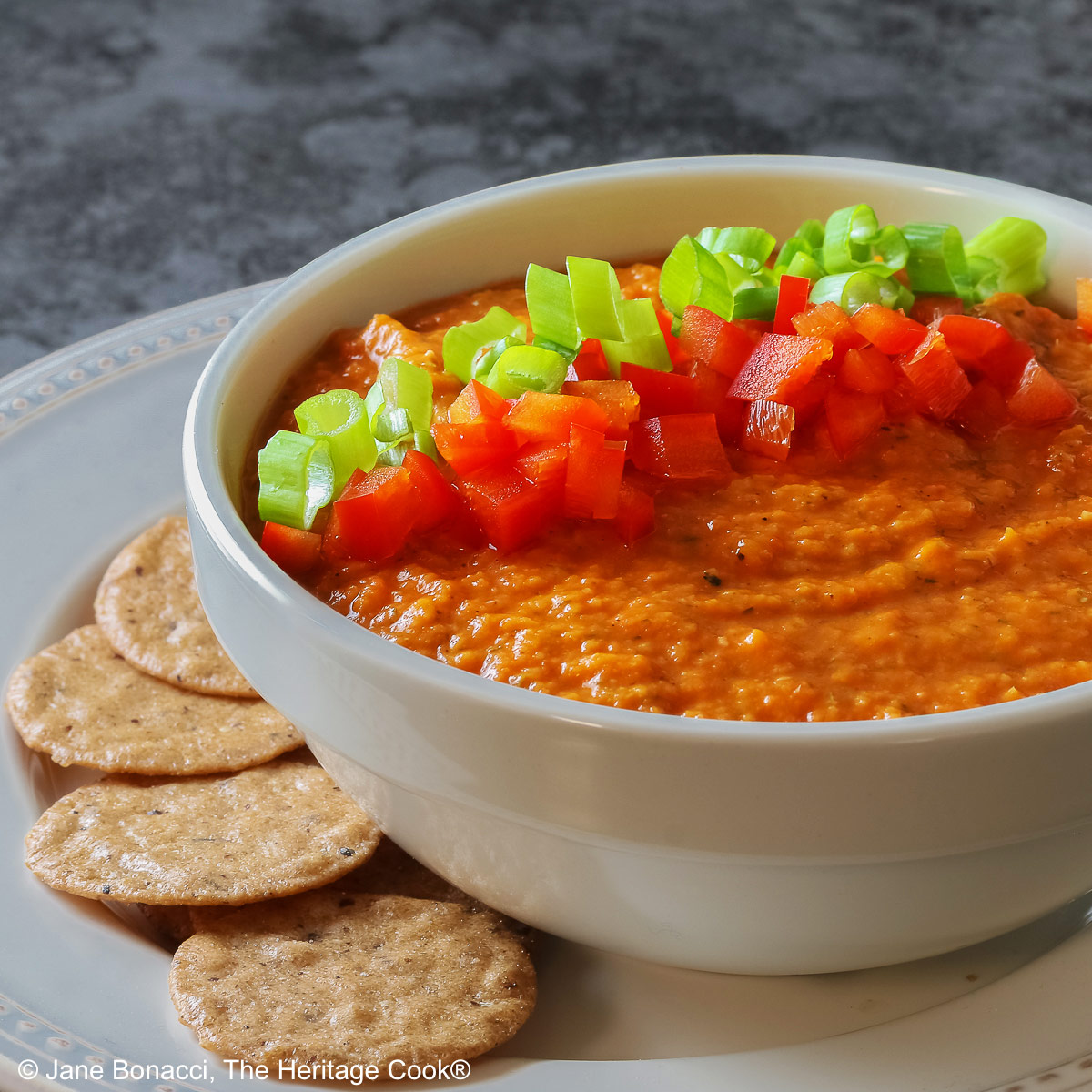 A white bowl full of White Bean and Red Bell Pepper Dip, topped with some finely chopped red bell peppers and finely sliced green onions, served with a ring of gluten-free Nut-Thin Pecan Crackers for dipping. The bowl and crackers are sitting on a cream colored plate with a red bell pepper in the back © 2026 Jane Bonacci, The Heritage Cook.