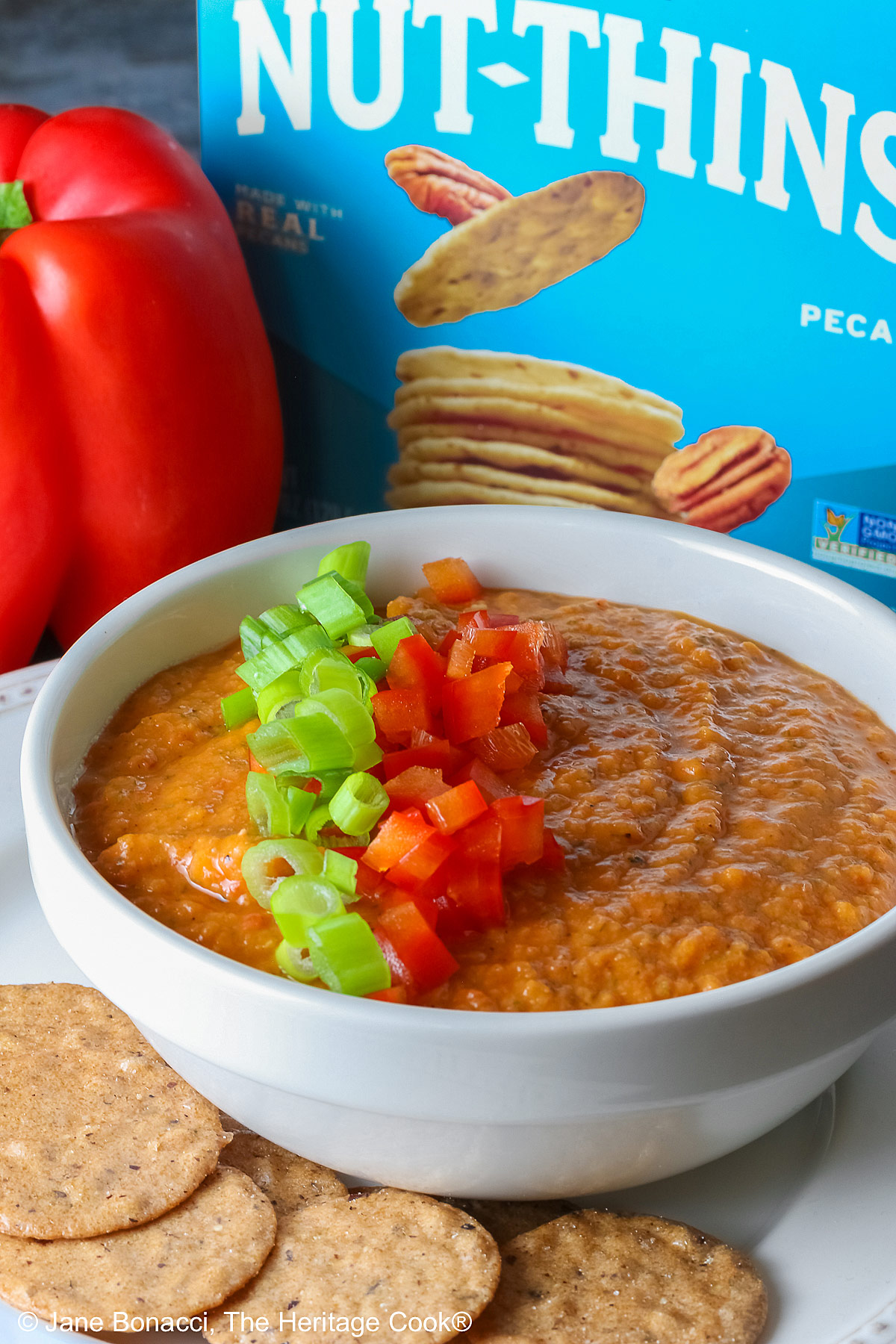 A white bowl full of White Bean and Red Bell Pepper Dip, topped with some finely chopped red bell peppers and finely sliced green onions, served with a ring of gluten-free Nut-Thin Pecan Crackers for dipping. The bowl and crackers are sitting on a cream colored plate with a red bell pepper in the back © 2026 Jane Bonacci, The Heritage Cook.
