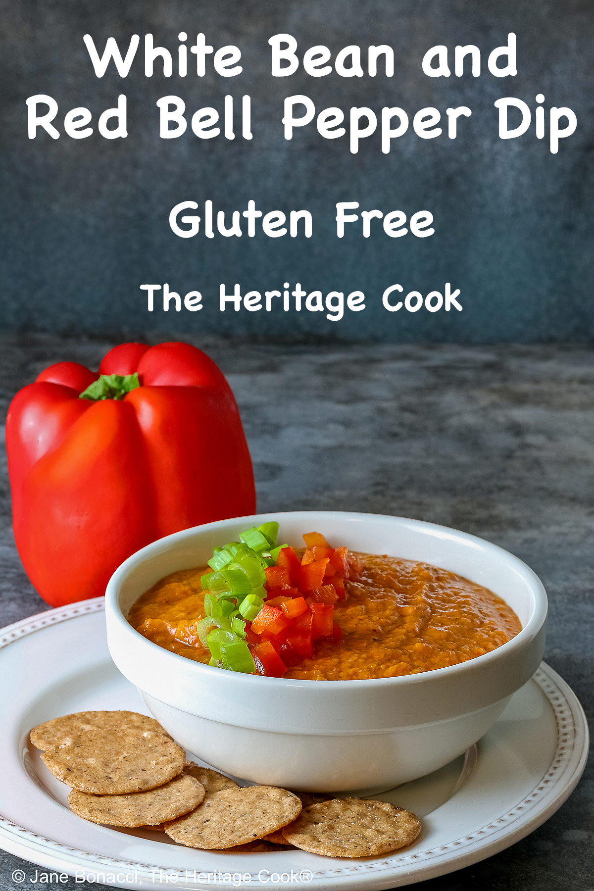 A white bowl full of White Bean and Red Bell Pepper Dip, topped with some finely chopped red bell peppers and finely sliced green onions, served with a ring of gluten-free Nut-Thin Pecan Crackers for dipping. The bowl and crackers are sitting on a cream colored plate with a red bell pepper in the back © 2026 Jane Bonacci, The Heritage Cook.