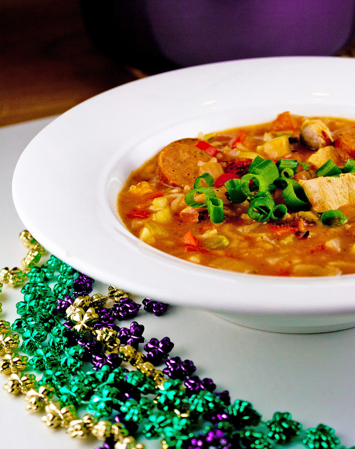 Hearty stew with chicken and sausage, flavored with creole seasonings and lots of vegetables, decorated with Mardi Gras beads alongside; Chicken Sausage Gumbo © 2023 Jane Bonacci, The Heritage Cook.