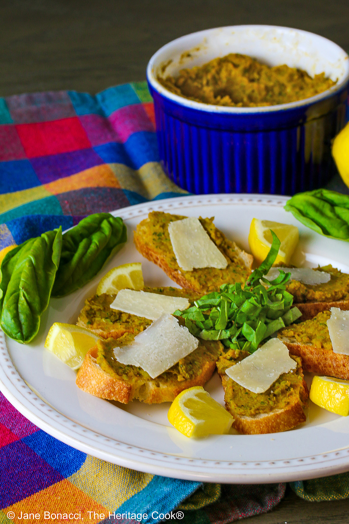 Fava bean spread on sliced baguette crostini with a shave of Parmesan on top and mini wedges of lemon beside each one, garnished with basil chiffonade and whole leaves on the white plate, with a whole lemon and bowl of the fava bean spread behind, all set on a brightly colored plaid cloth © 2025 Jane Bonacci, The Heritage Cook. 