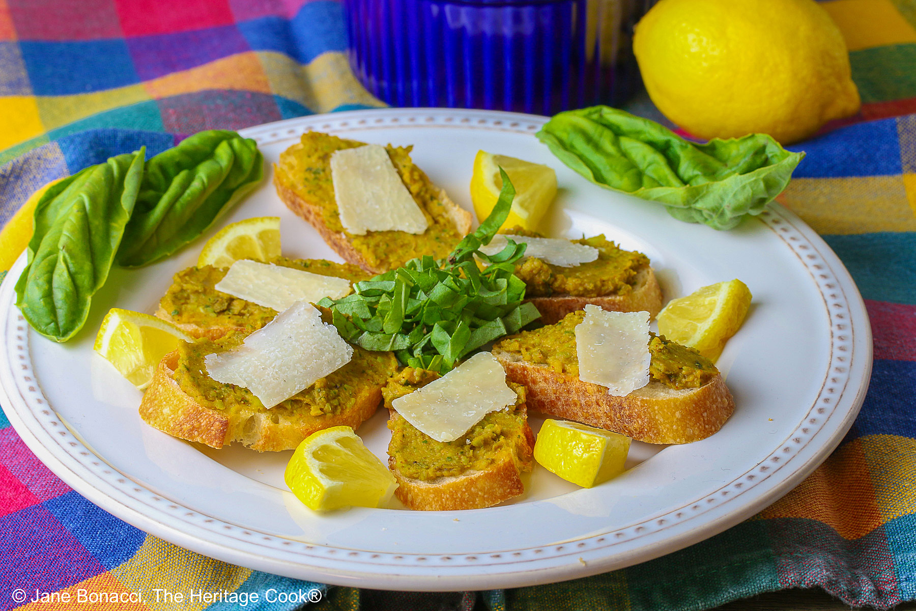 Fava bean spread on sliced baguette crostini with a shave of Parmesan on top and mini wedges of lemon beside each one, garnished with basil chiffonade and whole leaves on the white plate, with a whole lemon and bowl of the fava bean spread behind, all set on a brightly colored plaid cloth © 2025 Jane Bonacci, The Heritage Cook. 