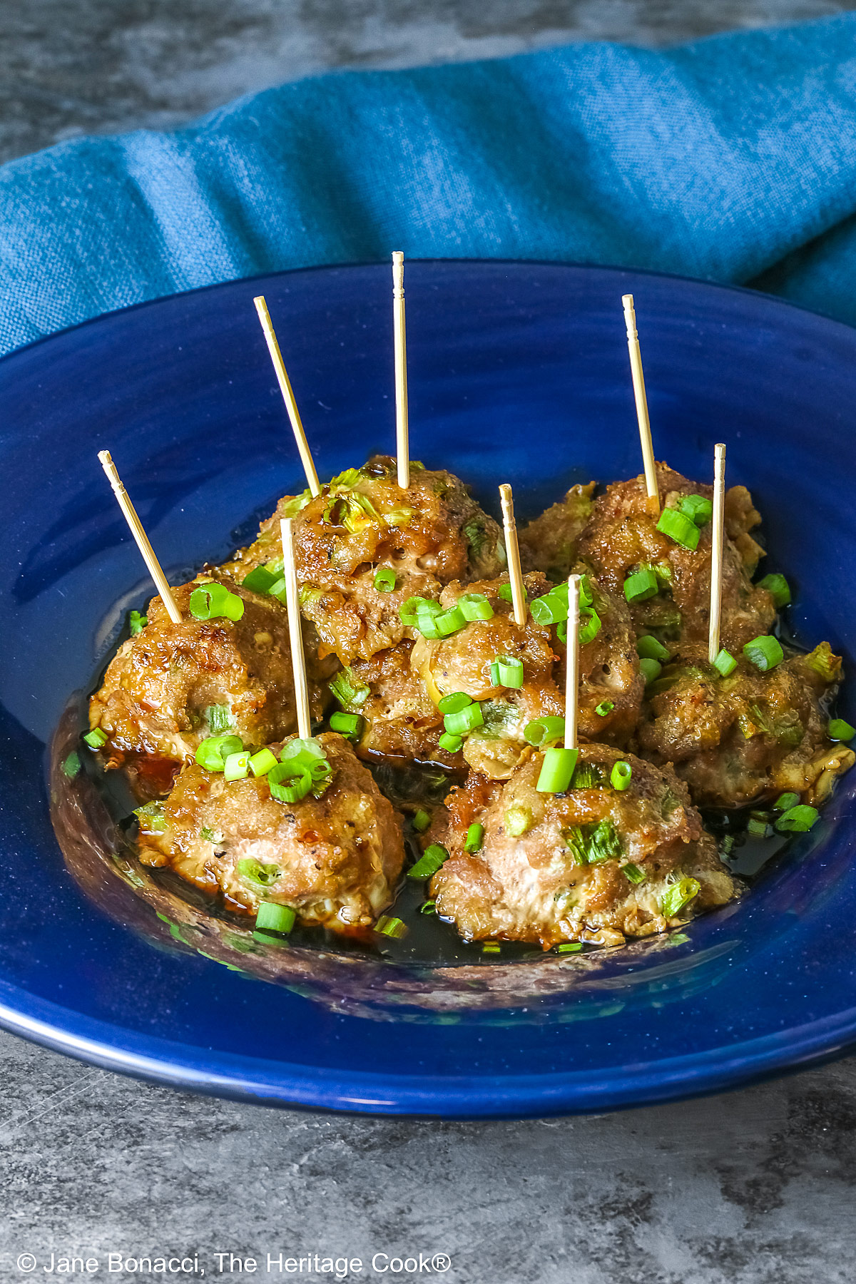 A piled stack of Turkey-Scallion Meatballs with Soy Ginger Glaze in a deep blue bowl with a wide rim and a lighter blue cloth behind, on top of a mottled gray background © 2026 Jane Bonacci, The Heritage Cook.