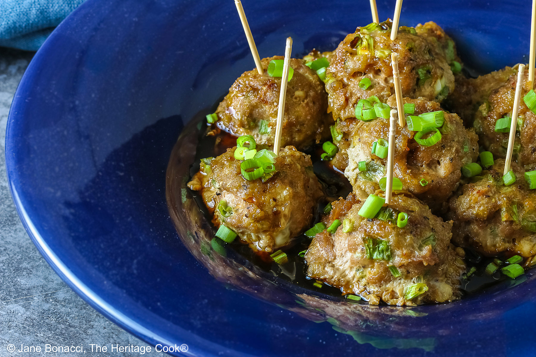 A piled stack of Turkey-Scallion Meatballs with Soy Ginger Glaze in a deep blue bowl with a wide rim and a lighter blue cloth behind, on top of a mottled gray background © 2026 Jane Bonacci, The Heritage Cook.