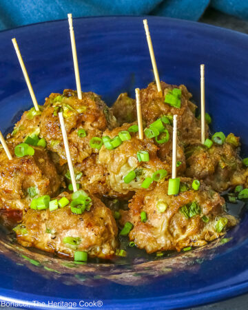 A piled stack of Turkey-Scallion Meatballs with Soy Ginger Glaze in a deep blue bowl with a wide rim and a lighter blue cloth behind, on top of a mottled gray background © 2026 Jane Bonacci, The Heritage Cook.