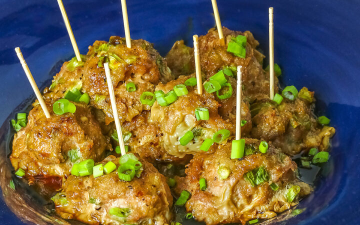 A piled stack of Turkey-Scallion Meatballs with Soy Ginger Glaze in a deep blue bowl with a wide rim and a lighter blue cloth behind, on top of a mottled gray background © 2026 Jane Bonacci, The Heritage Cook.