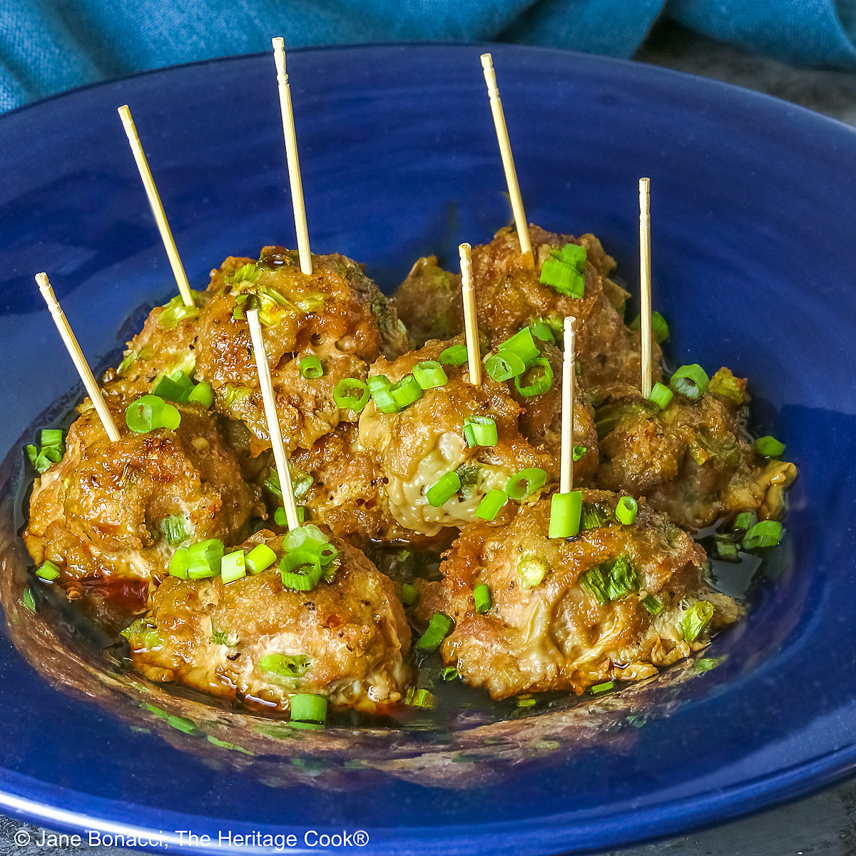 A piled stack of Turkey-Scallion Meatballs with Soy Ginger Glaze in a deep blue bowl with a wide rim and a lighter blue cloth behind, on top of a mottled gray background © 2026 Jane Bonacci, The Heritage Cook.