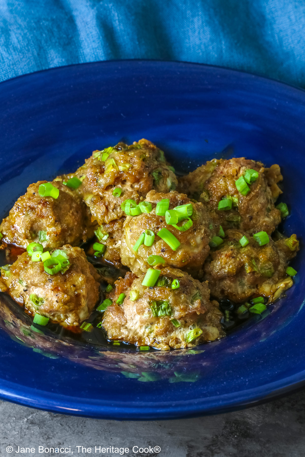 A piled stack of Turkey-Scallion Meatballs with Soy Ginger Glaze in a deep blue bowl with a wide rim and a lighter blue cloth behind, on top of a mottled gray background © 2026 Jane Bonacci, The Heritage Cook.