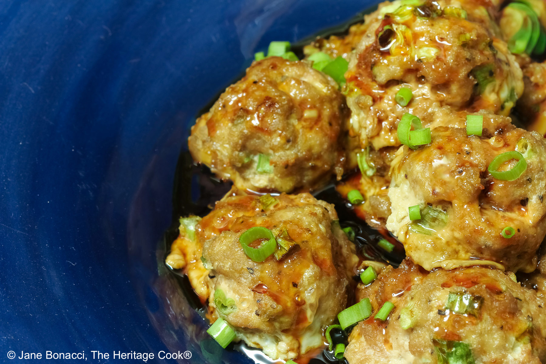 A piled stack of Turkey-Scallion Meatballs with Soy Ginger Glaze in a deep blue bowl with a wide rim and a lighter blue cloth behind, on top of a mottled gray background © 2026 Jane Bonacci, The Heritage Cook.