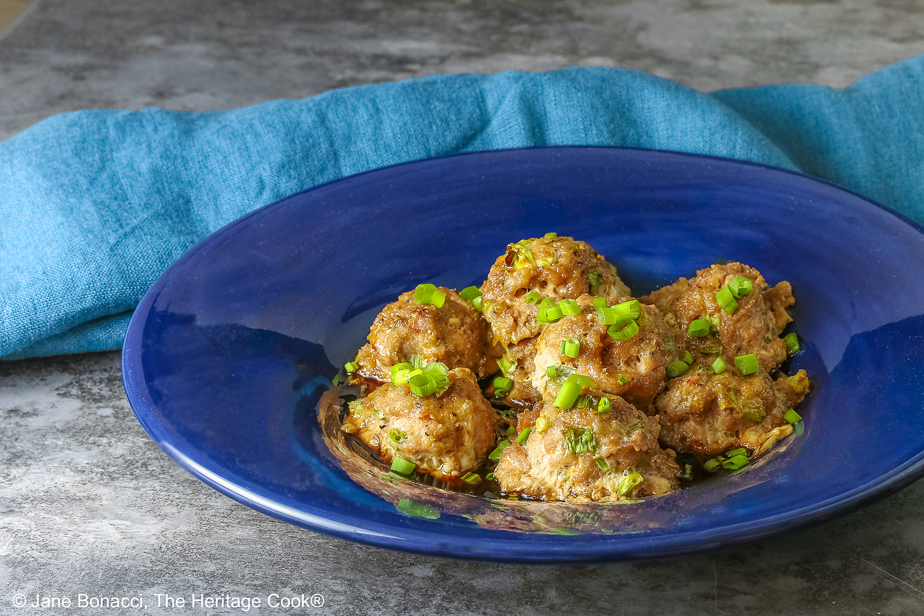 A piled stack of Turkey-Scallion Meatballs with Soy Ginger Glaze in a deep blue bowl with a wide rim and a lighter blue cloth behind, on top of a mottled gray background © 2026 Jane Bonacci, The Heritage Cook.