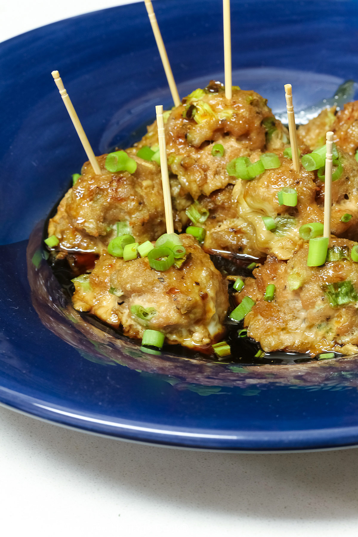 A piled stack of Turkey-Scallion Meatballs with Soy Ginger Glaze in a deep blue bowl with a wide rim and a lighter blue cloth behind, on top of a mottled gray background © 2026 Jane Bonacci, The Heritage Cook.