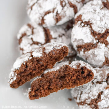 Luscious dark brown chocolate crinkle cookies covered with powdered sugar with cracks all over in beautiful patterns on a white plate or silver wire cooling rack some with a pitcher and glass of milk behind, on a black background © 2025 Jane Bonacci, The Heritage Cook.