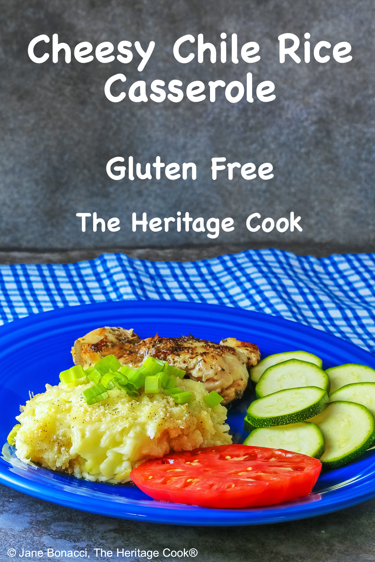 Bright blue plate filled with a piece of chicken, sliced zucchini, an heirloom tomato, large scoop of Cheesy Chile Rice Casserole topped with sliced green onions, with a blue checked cloth in the back and a fork in front © 2026 Jane Bonacci, The Heritage Cook.