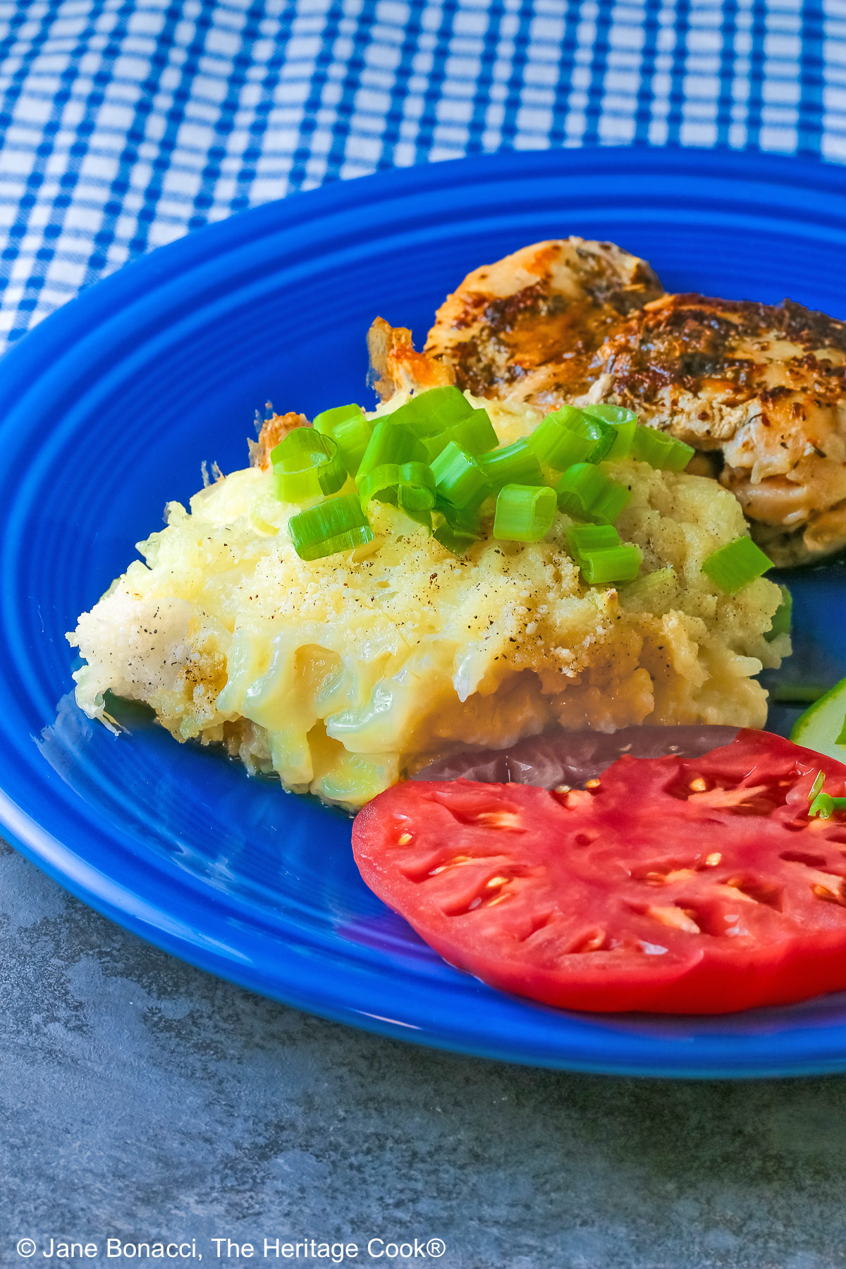 Bright blue plate filled with a piece of chicken, sliced zucchini, an heirloom tomato, large scoop of Cheesy Chile Rice Casserole topped with sliced green onions, with a blue checked cloth in the back and a fork in front © 2026 Jane Bonacci, The Heritage Cook.