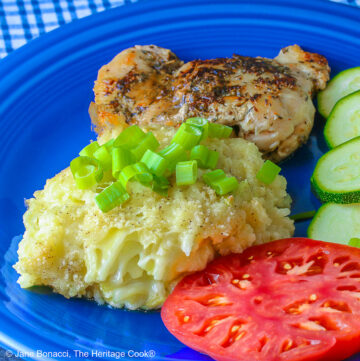 Bright blue plate filled with a piece of chicken, sliced zucchini, an heirloom tomato, large scoop of Cheesy Chile Rice Casserole topped with sliced green onions, with a blue checked cloth in the back and a fork in front © 2026 Jane Bonacci, The Heritage Cook.