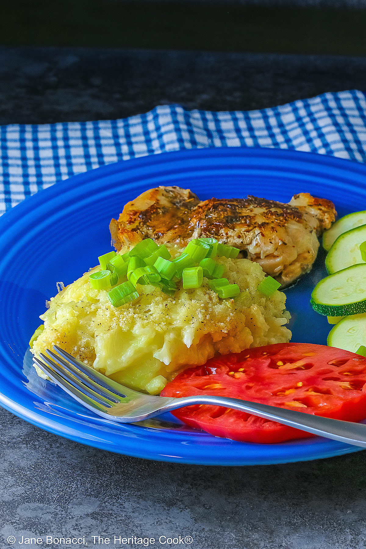 Bright blue plate filled with a piece of chicken, sliced zucchini, an heirloom tomato, large scoop of Cheesy Chile Rice Casserole topped with sliced green onions, with a blue checked cloth in the back and a fork in front © 2026 Jane Bonacci, The Heritage Cook.